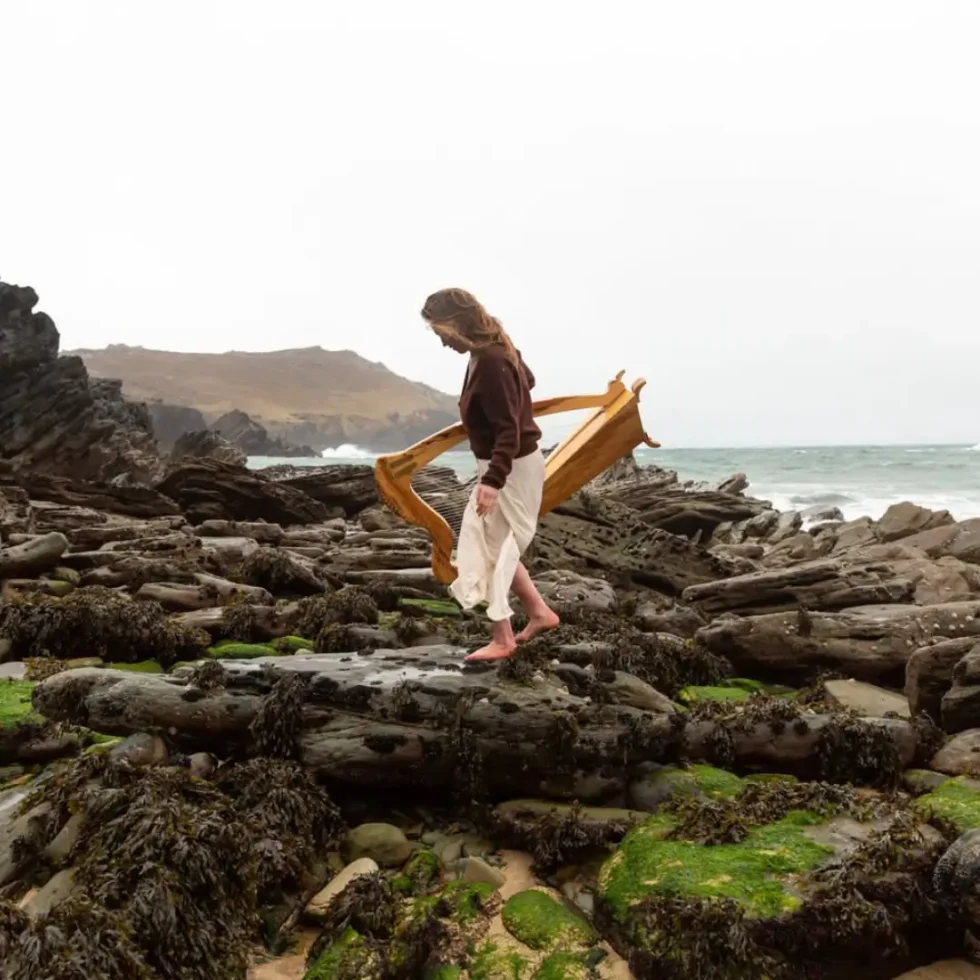 Cáit ni Riain on the rocky shore holding her harp and walking across the rocks in her bare feet