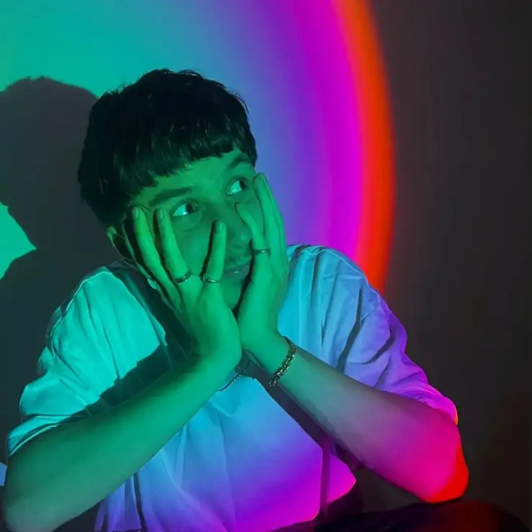 DJ Shee sitting in a multicoloured lit background with his hands ynder his chin